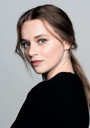 Eine junge Frau mit langen, blonden Haaren, die zu einem Zopf gebunden sind blickt über ihre linke Schulter in die Kamera. Sie trägt einen schwarzen Pullover