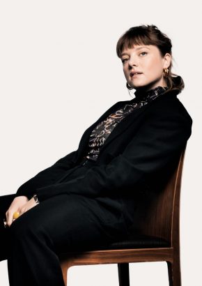 Eine junge Frau sitzt seitlich, angelehnt auf einem Holzstuhl und schaut mit neugierigem Blick in die Kamera. Sie trägt einen schwarzen Anzug und eine dunkle Bluse mit Muster