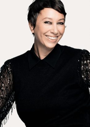Eine Frau mittleren Alters und schwarzen kurzen Haaren lächelt in die Kamera. Sie trägt eine schwarze Bluse mit transparenten und mit Pailletten bestickten Ärmeln