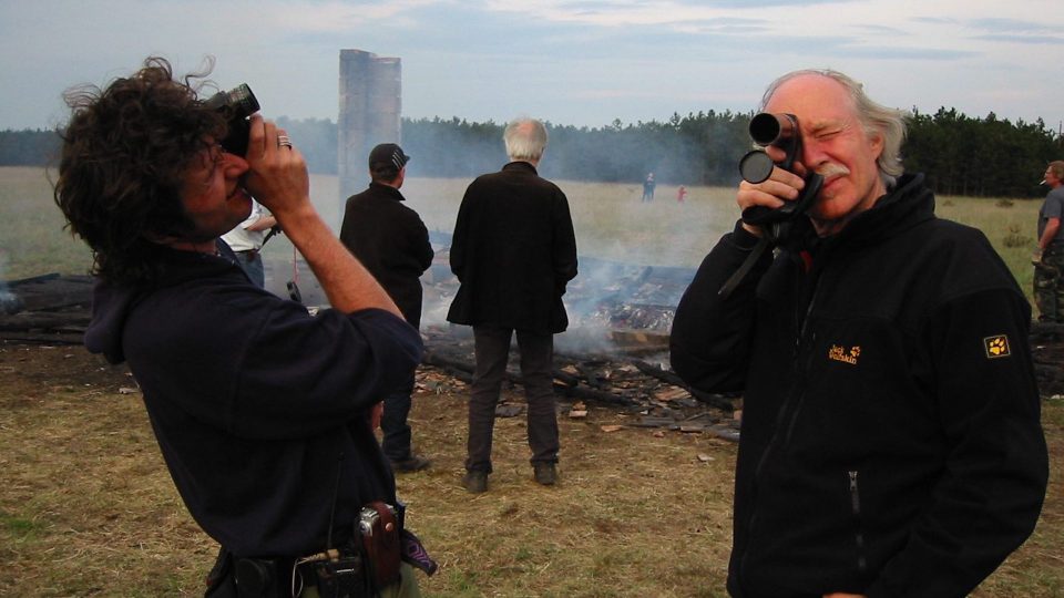Jürgen Jürges rechts, und eine Person von Crew links. Beide schauen jeweils durch ein Kameraobjektiv. Hinter ihnen eine brennende Ruine des Filmsets.