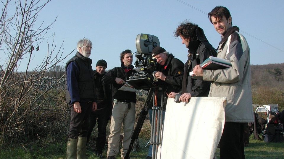 Jürgen Jürges mittig schaut in eine große Filmkamera. Michael Haneke in Gummistiefel und Crew stehen neben ihm.