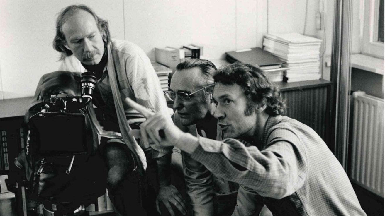 Schwarz-weiß Bild von Jürgen Jürges, Dennis Hopper und Roland Klick. Alle drei stehen um eine Filmkamera. Roland Klick zeigt mit dem Finger in eine Richtung.