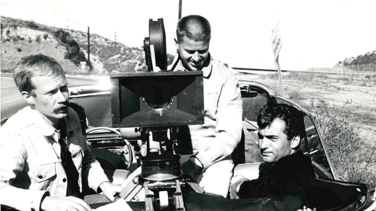 Schwarz-weiß Bild. Jürgen Jürges links, steht neben einer Kamera, die auf einem offene Cabrio-Auto angebracht ist. In dem Cabrio sitzen zwei Personen.