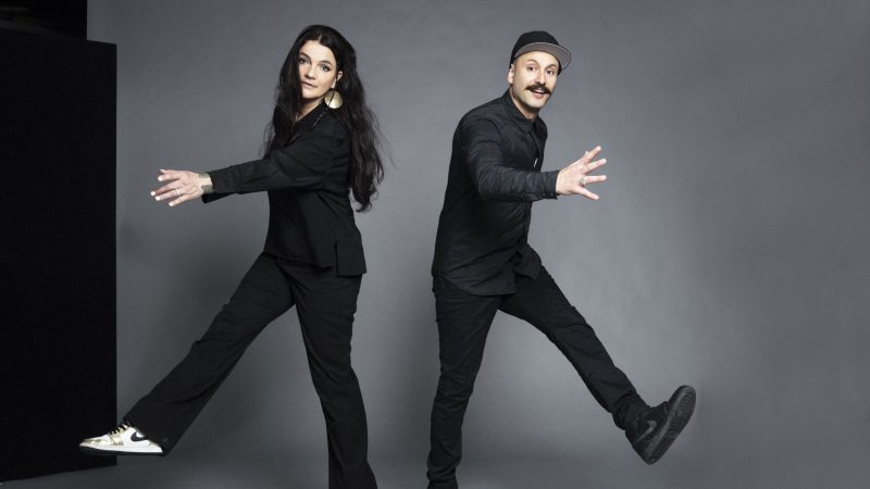 Regisseurin Katja von Garnier links und Choreagraf Vartan Basil rechts. Beide stehen nebeneinander vor einem grauen Hintergrund und haben jeweils einen Bein in der Luft und eine Hand gespreizt, als ob sie sich spiegeln würden.