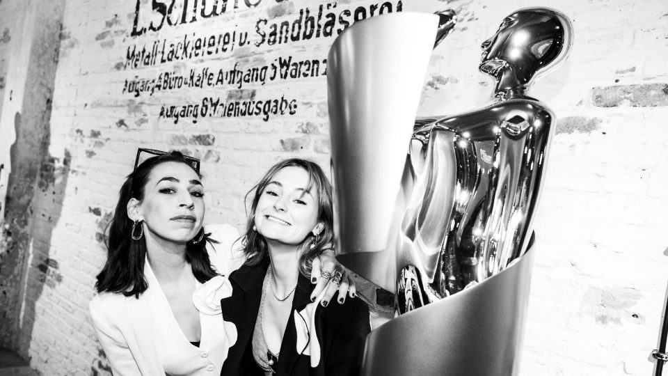 schwarz-weiß Bild: Carole Schuler & Lea van Acken stehen neben der großen Lolastatue, die Arme umeinander geschlagen.