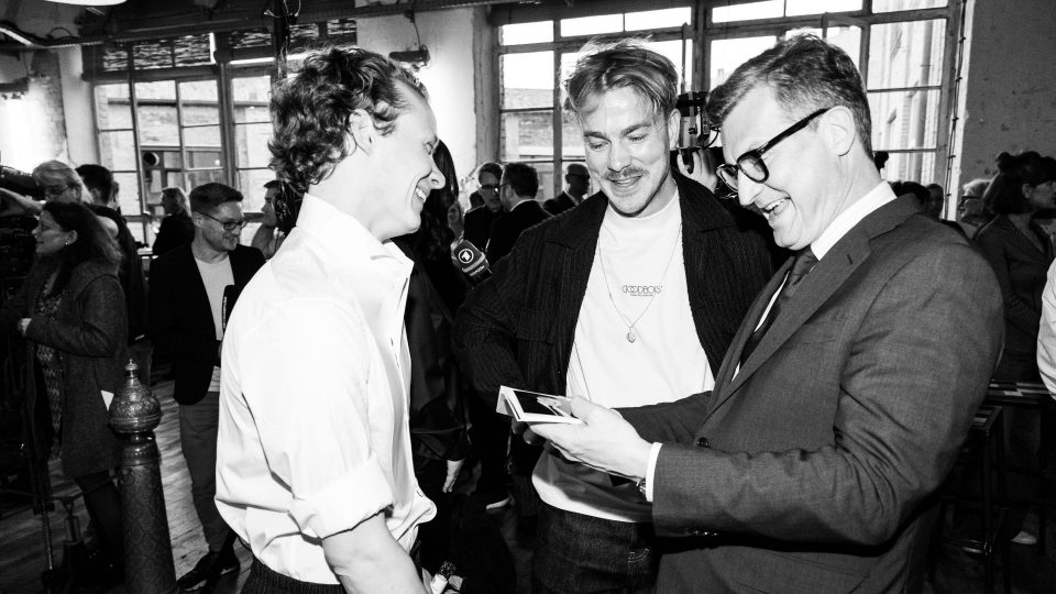 schwarz-weiß Bild: Felix Kammerer, Albrecht Schuch & Malte Grunert schauen sich lachend Polaroid-Bilder an