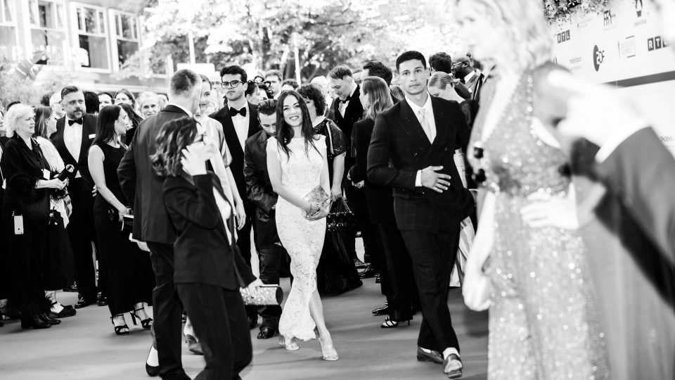 Mona Pirzat und Emilio Sakraya stehen im Fokus des schwarz-weiß Bildes. Um sie herum stehen viele Menschen in Kleidern und Anzügen.