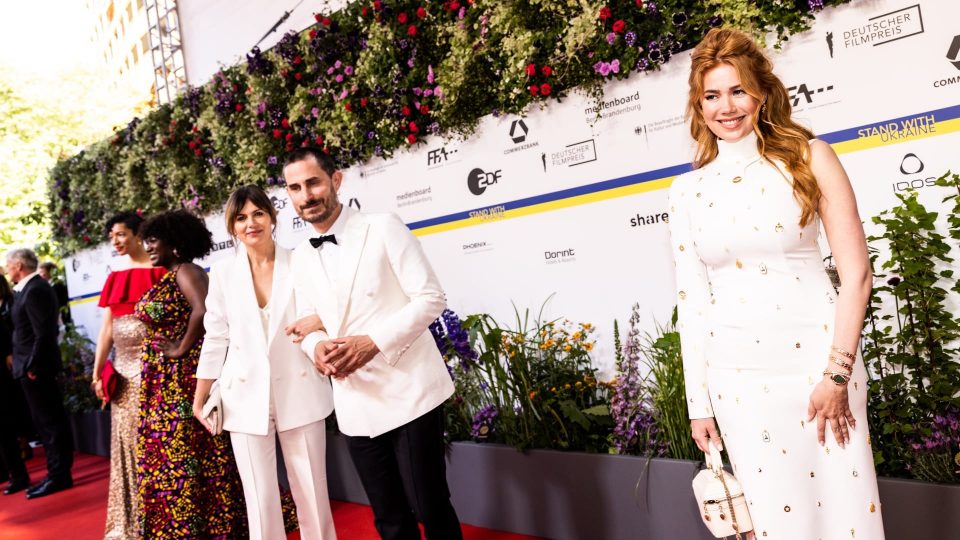Palina Rojinski trägt ein weißes Kleid mit goldenen Details. Sie lächelt. Im Hintergrund ist Clemens Schick mit einer Frau zusehen. Beide tragen weiße Anzüge.
