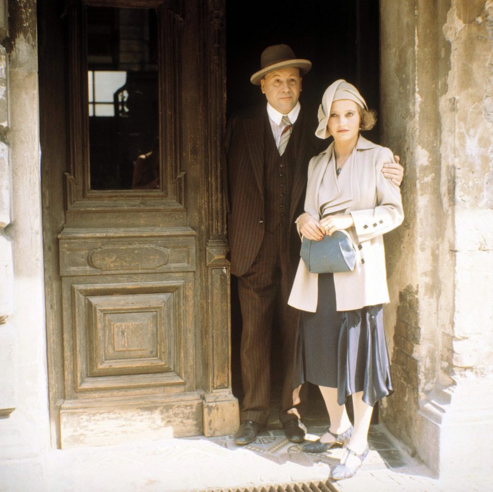 Hannah Schygulla und Günter Lamprecht stehen zusammen in einer braunen Tür. Er hat den Arm um sie gelegt und sie hält ihre blaue Tasche.