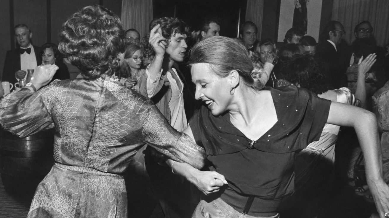 Hanna Schygulla tanzt mit einer anderen weißen Frau auf dem Eröffnungsempfang der 25.Berlinale. Die beiden haben ihren einen Arm verschenkt und drehen sich im Kreis.