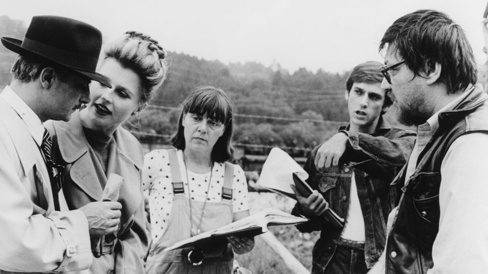 Zu sehen sind fünf weiße Personen, unter anderem Rainer Werner Fassbinder mit Hannah Schygulla. Das Bild zeigt eine Filmset, wo Anweisungen gegeben werden.