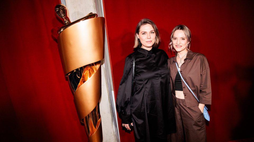 Cosima Vellenzer und Anika Klatt neben der LOLA-Statue. Cosima in einem schwarzen Blusenkleid, Anika in einem braunen Satin-Set aus Hemd und Hose mit einer hellblauen Abendtasche.