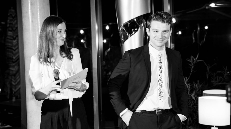 Das Schwarz-Weiß-Bild zeigt Alexandra Maria Lara neben Simon Morzé, der als Nominierter für die beste männliche Nebenrolle soeben auf die Bühne gerufen wurde. Sie trägt eine weiße Bluse, schwarze Hose und offene Haare, er einen schwarzen Anzug, ein weißes Hemd und eine Krawatte mit kleinen Grafiken.