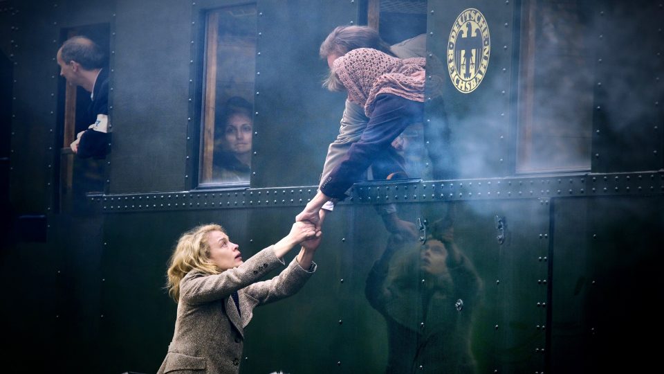Eine Frau und ein Mann lehnen aus dem Fenster eines anfahrenden Zuges, um die Hände einer Frau auf dem Bahnsteig festzuhalten. Sie schaut mit angestrengtem Blick zu den beiden hinauf. Neben dem Fenster prangt ein Emblem mit einem Adler und dem Schriftzug "Deutsche Reichsbahn".