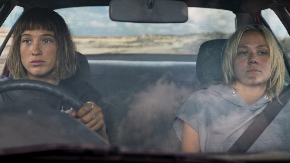 Im Auto: eine Frau mit schulterlangen dunkelblonden Haaren sitzt am Lenkrad, neben ihr eine mürrische Frau mit gebräunter Haut und hellblonden, stufigen Haaren in einem grauen Pullover.