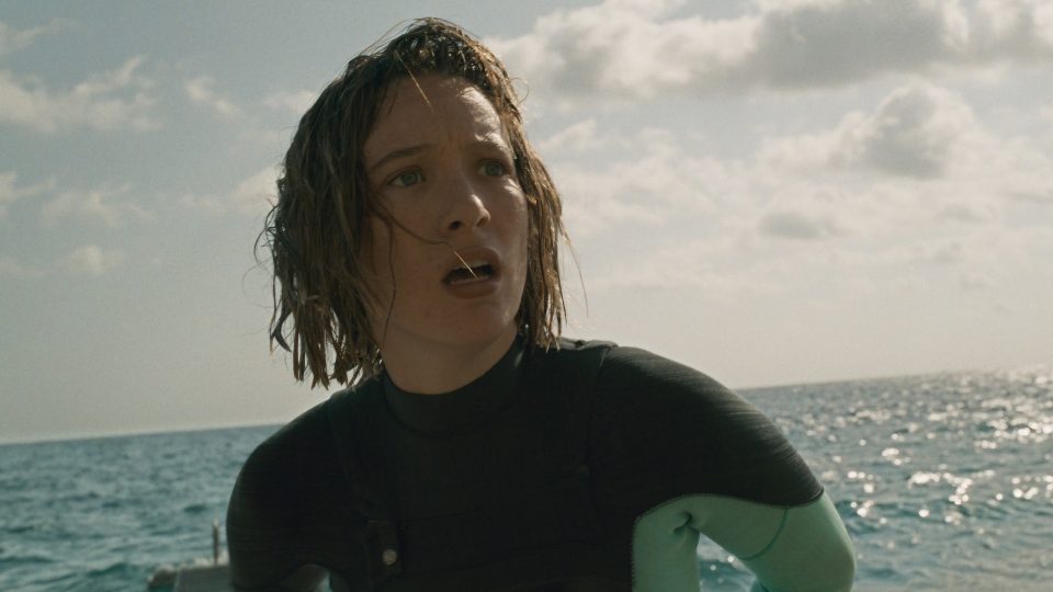 Eine Frau mit nassen, schulterlangen Haaren in Nahaufnahme. Sie trägt einen Taucheranzug, im Hintergrund liegt das glitzernde Meer. Ihr Gesichtsausdruck ist schockiert und besorgt.