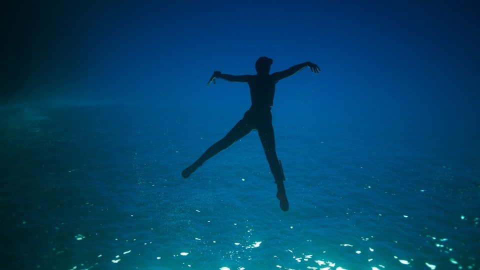 Unter der Meeresoberfläche: die Konturen eines Tauchers sind im Gegenlicht vor dem blauen Hintergrund zu erkennen, anmutig wie in einem Tanz.